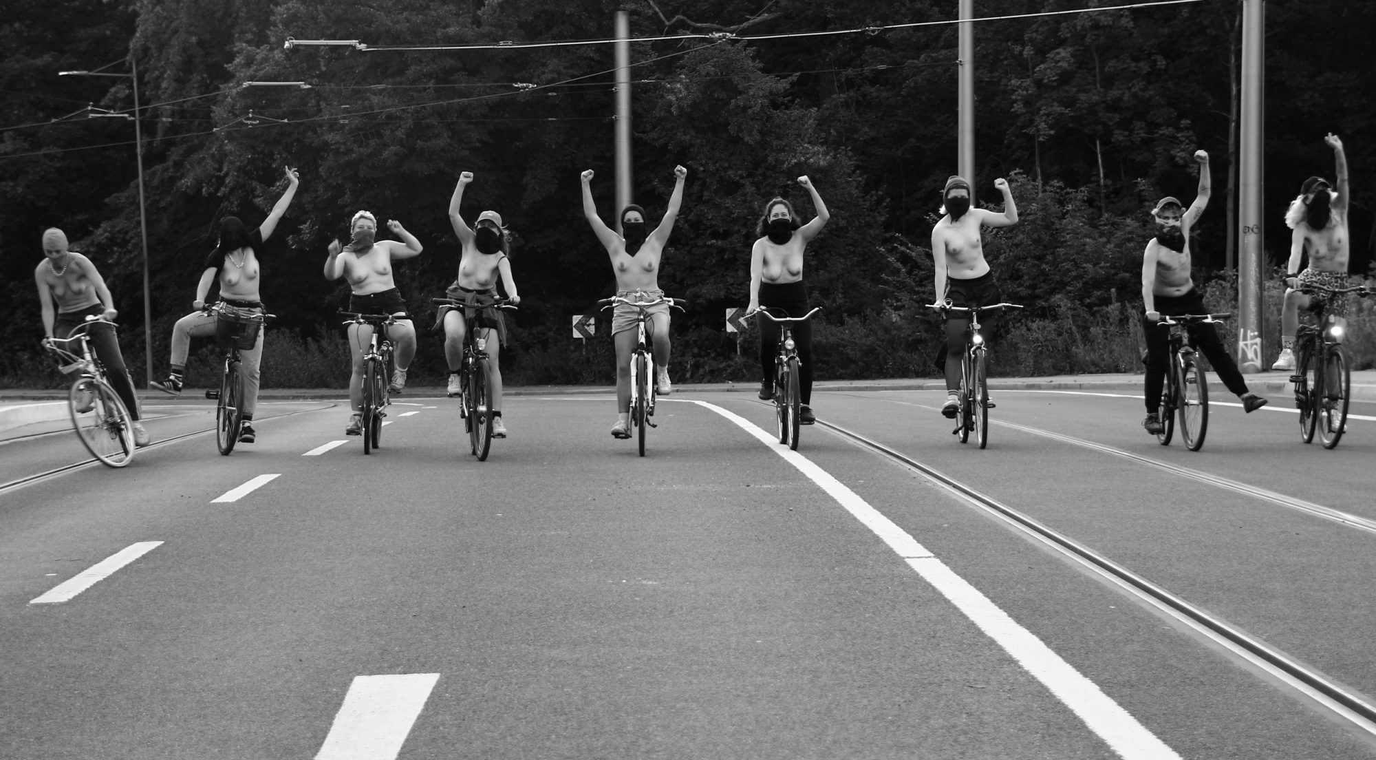 Schwarz-weiß Foto. 9 Personen sitzen auf dem Fahrrad. Oberkörperfrei und vermummt fahren sie auf Betrachter:in zu.